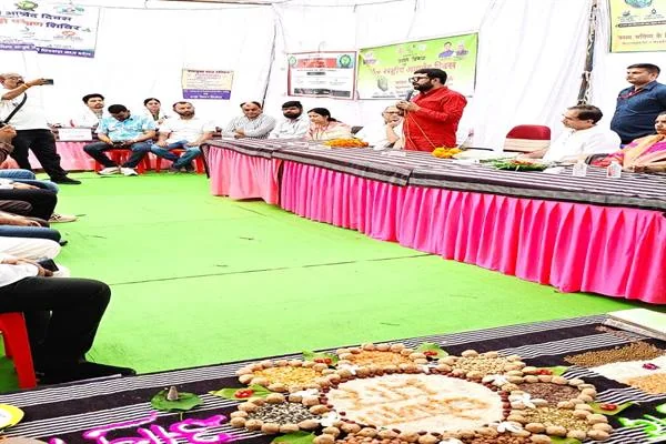 सभागार में मेज के पीछे बैठे अतिथियों के बीच माइक से बोलता एक व्यक्ति और सामने अनाज व प्राकृतिक सामग्री की सजाई गई प्रदर्शनी